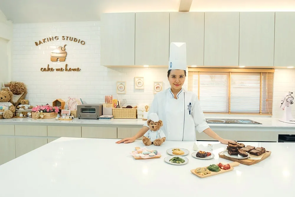เชฟทำขนมในครัวสตูดิโอ ใต้ป้ายโลโก้ BAKING STUDIO Cake Me Home